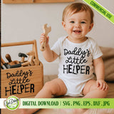 An adorable toddler sits on a wooden floor, holding a wooden toy wrench and smiling, next to a matching wooden toolbox that also has the 'Daddy's Little Helper' text and wrench-and-saw graphics applied. This heartwarming scene demonstrates how the SVG cut file can be used to create custom family-themed decor and apparel, appealing to crafters and DIY dads looking for a special Father's Day project.