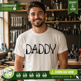A smiling man wearing a white t-shirt featuring the Daddy tools SVG graphic in a workshop, showcasing the perfect custom apparel for mechanic dads.