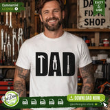 A smiling man with tattoos wearing a white custom t-shirt featuring the Handyman Dad SVG design with tool silhouettes in a rustic workshop setting, perfect for Father's Day apparel inspiration.