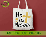 White tote bag featuring a black "He is Risen" typography and a golden cross design for Easter