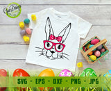 White kids t-shirt mockup printed with a black line art bunny face wearing pink glasses and a pink polka dot head bow on the chest; resting on a light grey wooden surface with tiny chicks, miniature eggs, and an Easter basket