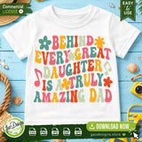 A trendy white t-shirt featuring colorful retro wavy typography that reads Behind Every Great Daughter Is A Truly Amazing Dad, styled on a blue wooden background with seashells to inspire summer Father's Day apparel projects.